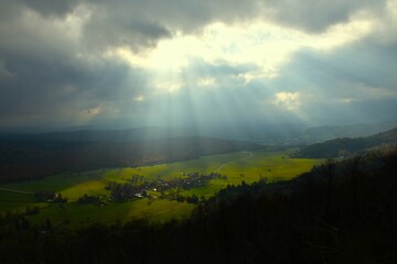 Obraz premium View of Strmca village at a field surrounded by forest on a cloudy day lit by sunlight in Notranjska, Slovenia