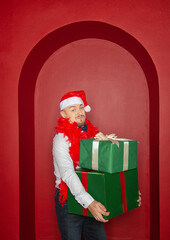 Young handsome smiling man wearing Santa hat holding Christmas gift boxes on the red arch background. New Year style
