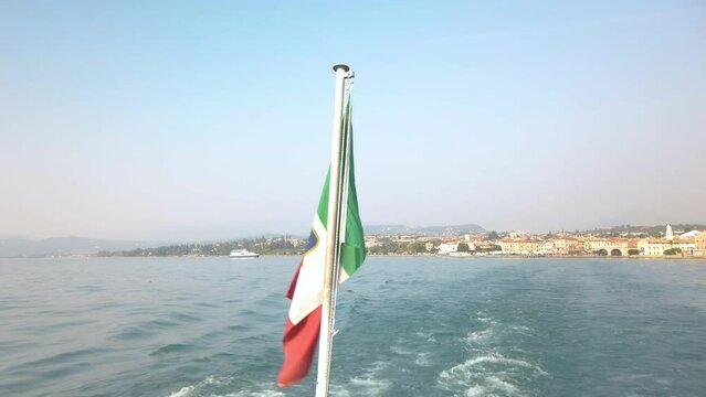 Flag waving gently in the wind onboard a tourist ferry on Lake Garda. The boat is leaving the port of Lazise, sailing towards Peschiera del Garda. It is a sunny summer day