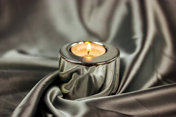 A burning candle in a metal candle holder on a table covered with a silver silk tablecloth....