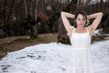 A woman in a white lace dress stands in a snowy forest, arms raised