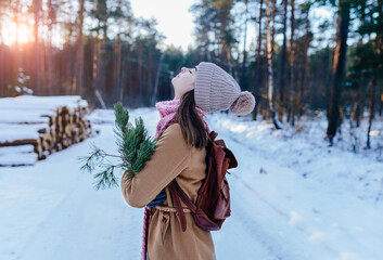 Beautiful brunette woman in brown coat with a winter bouquet of pine needles, spruce branches on the background of the winter forest. - obrazy, fototapety, plakaty