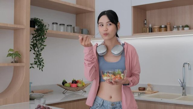 Smiling Asian pretty female eating salad with fruits and vegetables in kitchen