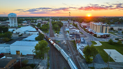 Golden sun setting on horizon with hotel and water tower split by train on railroad tracks aerial