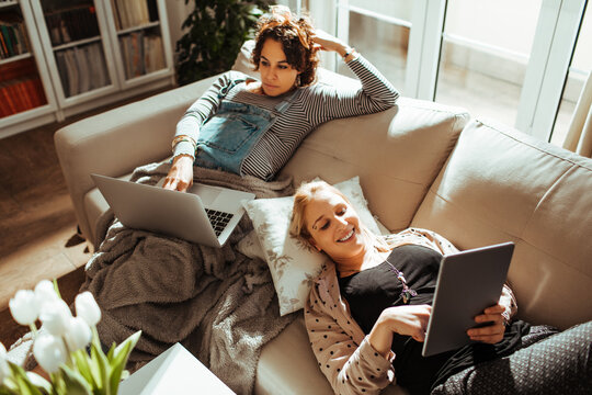 Two young women using gadgets on couch. Lesbian couple using laptop and tablet on the sofa at home
