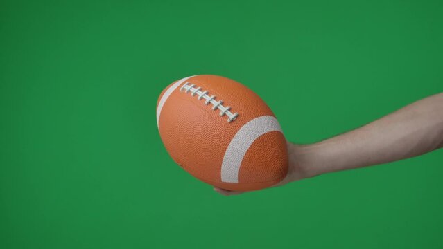 In a shot on a green background, chromakey male hand that holds, holds out an oval shaped ball for American soccer, it is inflated, bright color. This could be a place for your advertisement.
