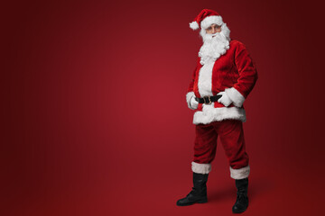 Jolly Santa Claus in traditional red suit standing with hands on hips against red backdrop