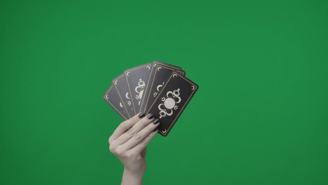 Framed on a green background, chromakey. A womans hand with a manicure holds playing tarot cards. She folded them like a fan and shows the outside with a beautiful pattern to the camera.