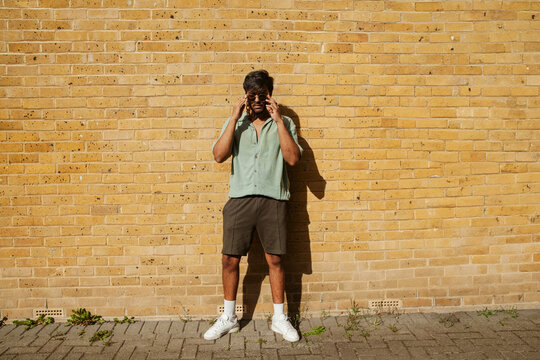Young Indian man in cool outfit and sunglasses leaning against brick wall