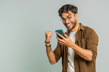 Happy excited Indian young man guy use smartphone typing browsing shouting say wow yes found out great big win good news lottery goal achievement celebrating success, winning play game on gray