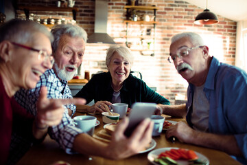 Senior man showing something funny on smartphone during lunch with friends at home