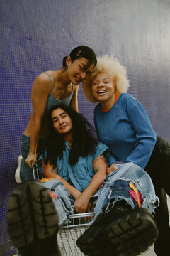 Two multiracial non-binary people and African woman with albinism sitting in shopping cart having fun outdoors