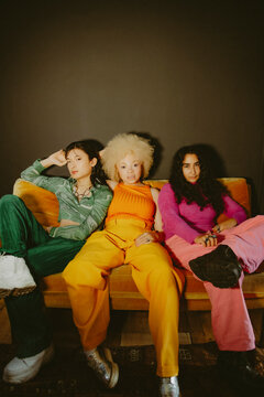 Three multiracial friends in bright fashionable outfits sitting on yellow couch at home