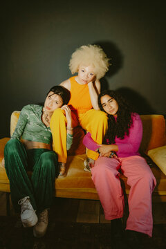 Three multiracial friends in bright fashionable outfits sitting on yellow couch at home looking into camera