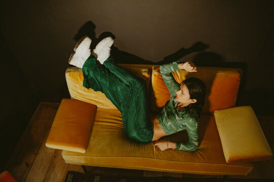 Young non-binary Asian person in fashionable green outfit posing on yellow couch in living room looking away from camera