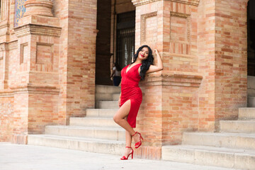 Fototapeta na wymiar Young and beautiful brunette and latin woman, dressed in short red dress and red shoes visits the famous square in seville, spain. The woman is leaning on a brick column and is happy. Travel concept.
