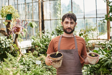 handsome and bearded gardener in linen apron smiling and holding plants in pots in greenhouse