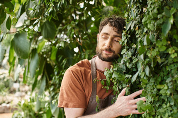 pleased bearded gardener in  linen apron smiling while working in greenhouse, horticulture concept