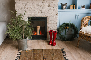 Spruce branches in a metal vase, red rubber boots and a Christmas wreath near the fireplace in the living room