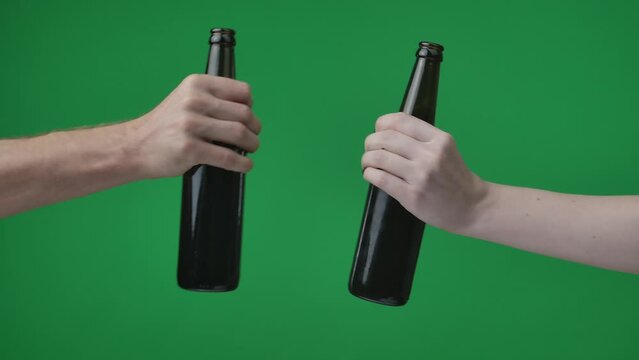 In a close up shot against a green background. A woman and a mans hand holding bottles of beer, alcohol and reaching out to each other to clink. They are drinking alcohol with an interlocutor