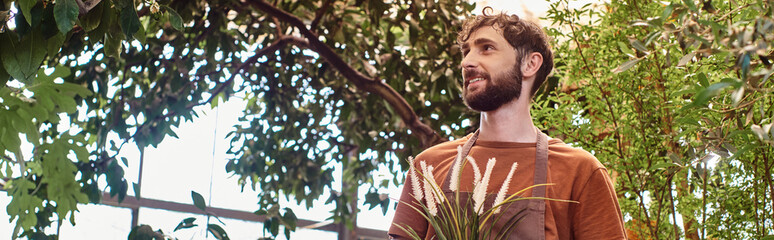 happy and good looking gardener in linen apron holding plant and standing in greenhouse, banner