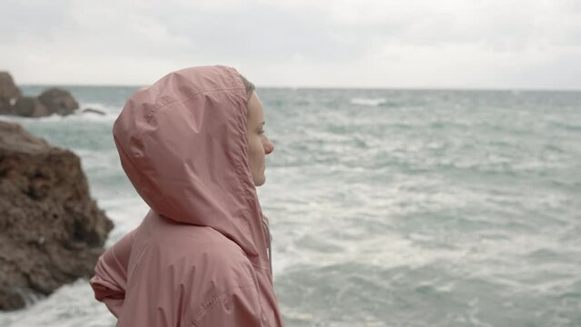 The camera captures sharp rocks and a young woman standing on them at the water's edge. There is a storm with strong waves at sea. She is wearing a raincoat.