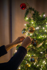 Female hands tying lights and baubles to Christmas tree indoor. Xmas preparations and tree decoration for the holiday season
