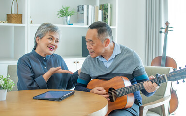Asian sweet senior elderly couple wearing casual clothes, staying in indoor cozy home at living room, playing guitar, singing together with happiness, smiling. Retirement, Healthcare Concept.
