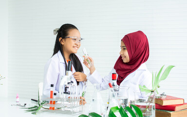 Two diverse multiethnic little girls wearing white ground uniform, hijab, headscarf, studying science in classroom at school, using stethoscope for research plants, happily smiling. Education Concept.