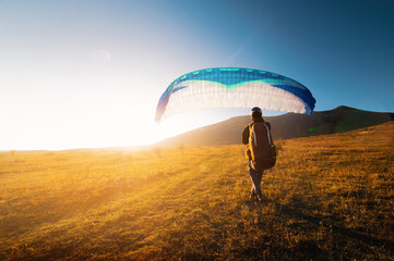 male paraglider takes off from a yellow field with a blue parachute against the backdrop of hills...