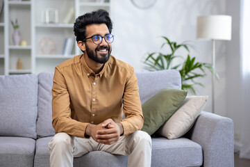 Successful young Indian businessman, freelancer sitting on sofa at home smiling and looking away.