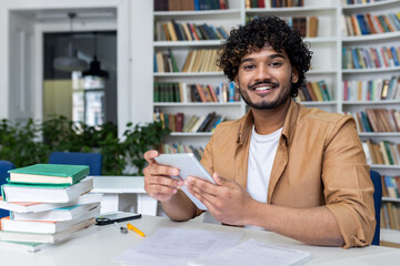 Portrait of a young Indian male student sitting in the university library, holding a tablet and...