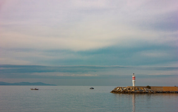 Seascape image with lighthouse on stone harbor, calm sea and cloudy sky. Lighthouse and small boats on the sea cinemagraphic view, landscape image.
