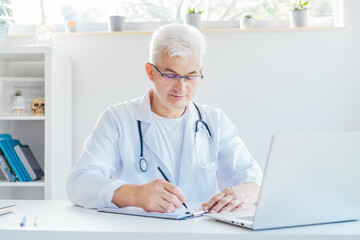Confident mature caucasian senior male doctor making notes while using laptop for telemedicine with patients at hospital. GP make online video call consult patient. Online remote medical appointment.