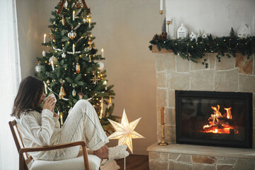 Beautiful woman in stylish pajamas with warm tea relaxing at decorated tree and fireplace, enjoying cozy christmas morning in festive living room with gifts. Merry Christmas! Winter hygge