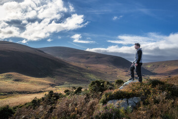Adult man standing on the rock, illuminated by sunlight, and looking at distant mountains. Hiking in Wicklow Mountains, Ireland