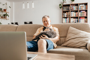 A man with a gray cat in his arms sits on the sofa and watches a program, television through a...