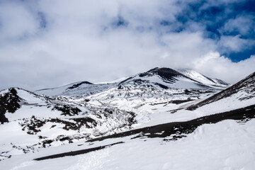 Crater of Mount Etna volcano in winter, Sicily island, Italy. Landscape of Silvestri craters with black volcanic lava stones. Active volcano slopes covered with snow under blue sky, clouds and smoke