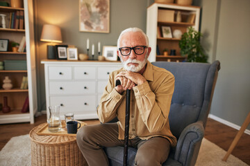 Old man holding hands on wooden stick, resting in cozy armchair in living room