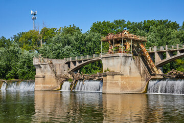 Rusty Maumee River Dam cogs and equipment with logs over waterfalls into basin and green trees Fort Wayne, IN