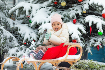 Cute pretty little girl outdoor sitting in a sled on snow near decorated snow-covered Christmas tree, during snowfall, in forest, near old wooden house. happy time on snowy