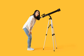 Young Asian woman looking at stars through telescope on yellow background