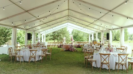 Wide shot of beautiful decorated tables for the wedding under the tent. Bouquets of wild flowers in pink colors are on the festive tables, wine glasses and plates, on outdoor wedding, slow motion.