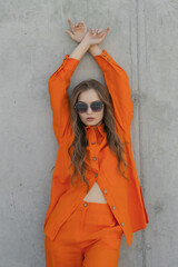 young lady in sunglasses and orange pantsuit at concrete wall
