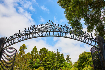 The Gardens at Lake Merritt gate arch entrance on blue sky day with clouds over green treetops