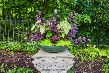 Fototapeta na wymiar Plants in blue bowl, planter atop white Greek pillar with mulch leading to black fence and forest