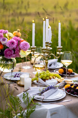 Romantic sunset wedding table decor for a couple with vintage chandelier, burning candles, snacks, white wine, fruits, ranunculi flowers. Summer vibes, field or meadow, outdoors event