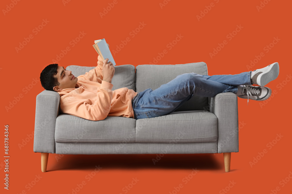 Poster handsome young man reading book on grey sofa against orange background