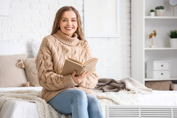 Young woman reading book near radiator in bedroom