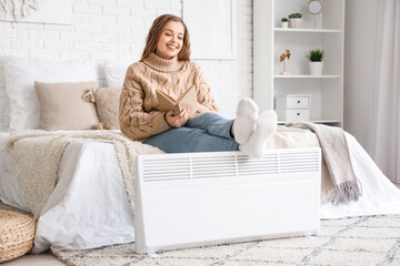 Young woman with book warming legs on radiator in bedroom - obrazy, fototapety, plakaty